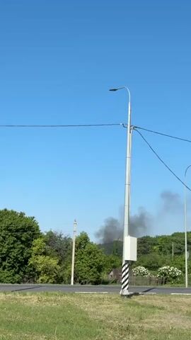 The video shows a rural scene with a tall, silver utility pole and street lamp in the foreground. The pole has a small, white box attached and is painted with black and white stripes at its base. Black power lines stretch overhead. To the left and right of the pole are patches of green grass and sections of a paved road. In the background, beyond the road and grass, there is dense greenery with trees and shrubs, and a small building can be glimpsed amongst the trees. Rising behind the trees is a plume of thick, black smoke obscuring part of the clear blue sky.