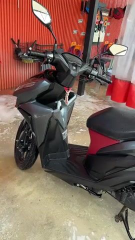 The video shows a gray Honda Click motorcycle being washed. The motorcycle is parked in what appears to be a car wash. There's a red-trimmed white curtain partially visible in the background, along with washing tools.  Foam is present on the ground around the motorcycle. A black pickup truck is partially visible in the background behind the motorcycle.  The camera pans around the motorcycle, showing its front, side, and rear.