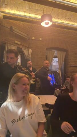 The video captures a lively scene inside what appears to be a restaurant or bar. The setting features exposed brick walls, warm ambient lighting from overhead fixtures, and ceiling-mounted heaters.

In the foreground, a young woman with blonde hair, wearing a white t-shirt with "Balenciaga" written on it, is seated at a table. She is smiling broadly, moving her hands to the music, and at one point, she claps her hands. To her right, another woman with dark hair is partly visible, also smiling and moving her hands in enjoyment.

In the background, a band is performing. On the far left, a man in a black shirt is playing a violin. Behind him, another man is seen playing a string instrument, possibly a guitar. The central figure of the band, to the right, is a man playing an accordion that features blue illuminated accents. Other band members or patrons are visible further in the background, some moving around.

The atmosphere appears festive and enjoyable, with the visible patrons clearly engaged with the live music. The tables are covered with checkered tablecloths. Through a window in the background, part of a sign that reads "osa" is visible.
