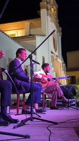 A group of musicians perform on a stage outdoors at night. The stage is made of wooden planks and is lit with purple lighting. In the foreground, the legs of two men are visible; one man on the left is wearing black dress shoes and dark pants, and another man to his right is wearing dress shoes and dark pants. In the middle ground, two men are seated on chairs performing. The man on the left wears a dark suit jacket and pants and is singing into a microphone. He has dark hair and is looking up. The man on the right wears a white jacket and light-colored pants and is playing a guitar. He has dark, curly hair and is looking down at his guitar. Two microphones with stands are positioned in front of the performers. In the background, a white building with a bell tower is visible. The building is lit from above.