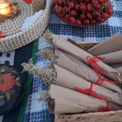 A still shot shows a picnic spread on a blue and white checkered blanket. In the foreground, several bunches of dried grass or flowers are wrapped in brown paper and tied with red string. To the left, a wicker basket holds more dried decorative items, including what appears to be a pinecone-shaped candle and an orange artificial flower. Behind the basket, a glass bowl overflows with ripe red cherries. To the right, a clear plastic container holds sliced red apples. In the center of the frame, a wicker tray is decorated with artificial flowers in red, orange, and white, a lit tea light candle in a glass holder, and a pinecone-shaped candle. The lighting suggests it is either early morning or late afternoon.