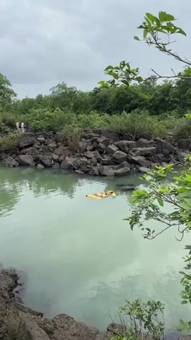 A still, pale green body of water is surrounded by dark, jagged rocks. In the distance, a dense forest of green trees forms a lush backdrop. A colorful, striped raft floats horizontally in the water. Several white birds are perched on the rocks. In the upper left of the frame, three individuals stand on the rocks. The sky is overcast and grey.