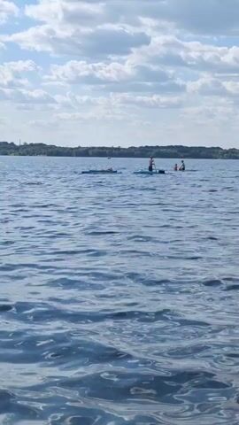A group of people are on a lake. Two people are on paddleboards. One person is standing on a paddleboard and appears to be paddling. Another person is sitting on a paddleboard. A third person is lying down on a paddleboard. The lake is calm with small waves. The sky is blue with scattered clouds. There is a tree line in the background.