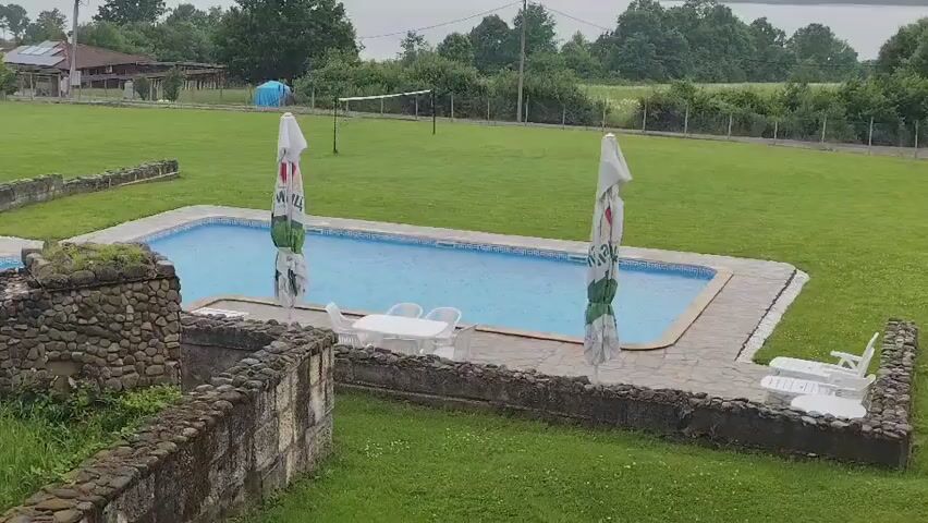 An outdoor, static, high-angle shot captures an empty recreational area under an overcast sky. In the center, a rectangular swimming pool with blue water is flanked by two large, colorful beach umbrellas. A white plastic table with four matching chairs sits beside the pool, and two white lounge chairs are visible on the right. The pool area is paved with flagstones and surrounded by a vast expanse of green grass. In the foreground, low stone walls or planters delineate the space. The background features a volleyball net on the grass, a fence, dense trees, and a building with a red roof and solar panels on the far left. No people are present.