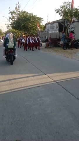 A group of children in red and white uniforms are marching in formation down a paved road. They appear to be participating in a parade or similar event. Several adults are observing from the side of the road, some on motorcycles. The environment is outdoors, with buildings and trees visible in the background. The sky is bright, suggesting it is daytime.