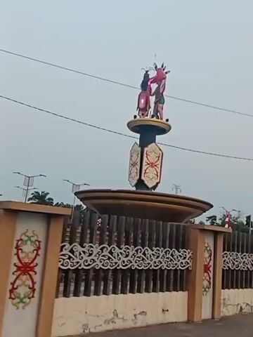 The video shows a static shot of an outdoor monument with decorative fencing in the foreground. The monument features two figures on top, dressed in traditional attire. Below them, a cylindrical section of the monument displays ornate panels with what appears to be a tribal pattern. The monument is situated within a wider landscape that includes flags, a traffic light, and power lines, suggesting an urban or semi-urban setting. The sky is overcast, and the overall lighting suggests it is daytime. The camera appears to be stationary, capturing the monument from a slightly low angle.