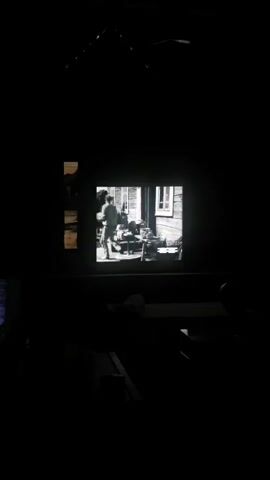 A black and white video is projected onto a screen in a dark room. The video shows a group of people, some of whom are seated around a table outdoors. There appears to be a wooden building with windows behind them. The people are dressed in what looks like historical clothing. The projection is centered and appears to be the main focus of the scene.