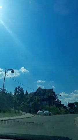 The video shows a view from inside a car driving on a road. The sky is clear and blue with some scattered clouds. There are houses and trees along the side of the road, and the car passes by a white car and then a black car. The road appears to be in a hilly or mountainous area. The video is short and appears to be a quick snapshot of a drive.