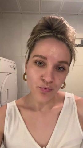 A woman stands in a bathroom, facing the camera. She has blonde hair in a high bun, and is wearing a white tank top and gold hoop earrings. She also has a small ring through her right nostril. Behind her is a white tiled wall, a white washing machine, and a chrome towel rack. She makes a kissing face and then shakes her head. Upbeat music plays in the background.