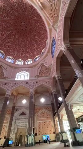 The video shows the interior of a large, ornate room, likely a mosque, with a massive pink dome dominating the view. The dome is intricately decorated with geometric patterns in a lighter pink hue. Numerous arched windows, some with blue panes, are visible around the base of the dome and along the upper walls. Large, thick columns, appearing to be a mix of dark brown and pink, support the ceiling and are spaced throughout the room. The walls are adorned with decorative panels, also in shades of pink and beige, featuring repeating motifs. The floor is covered with a patterned carpet, predominantly in shades of red and beige. Several tall, slim figures are sparsely scattered throughout the space, appearing to be people. Some are standing, while others might be walking. One prominent object in the foreground is a tall, white, cylindrical kiosk with a screen on its side. The lighting in the room appears to be a mix of natural light filtering through the windows and some artificial lighting, casting a soft glow throughout the space. The overall impression is one of grandeur and intricate architectural detail, with a sense of spaciousness and calm.