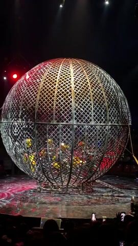 A crowd watches as several motorcyclists perform inside a large, spherical metal cage in a dimly lit arena. The cage, with a criss-cross patterned metal exterior, is rotating, allowing the motorcyclists to ride on the interior walls. Red and yellow lights illuminate the cage and the surrounding stage. The audience, visible in silhouette at the bottom of the frame, holds up cell phones to record the performance.