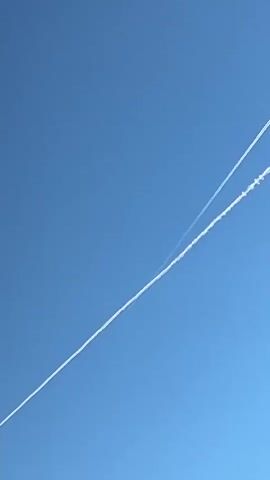 The video shows a clear blue sky with two contrails from airplanes. One contrail is a thin, unbroken white line. The other contrail is wider and appears to be breaking apart, with gaps in the white trail. The camera appears to be angled upwards towards the sky. There are no people or other objects visible in the frame.