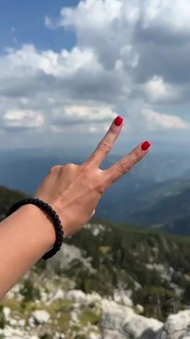The video shows a person's hand with red nail polish holding up two fingers in a peace sign against a backdrop of a cloudy sky and a vast mountain landscape. The hand is wearing a black beaded bracelet. The mountains are covered in trees, and there are rocky formations in the foreground. The sky is blue with scattered white clouds.