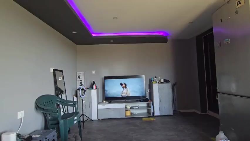 The video shows a room that appears to be a living area. The walls are painted a neutral grey, and the ceiling has a darker grey section with recessed lights and a strip of purple LED lighting around the edge. A flat-screen TV is the focal point, playing a video of a person wearing a virtual reality headset. Below the TV is a grey and white TV stand with items on the shelves. To the left of the TV is a small white cabinet with items on top, including a Christmas tree decoration, and a ring light on a tripod. To the right is another taller white cabinet. A green plastic chair sits on the left side of the room, near a mirror leaning against the wall. A refrigerator is visible on the right side of the room. A dark doorway or closet is also visible in the background. The floor is a grey tile. Music is playing.