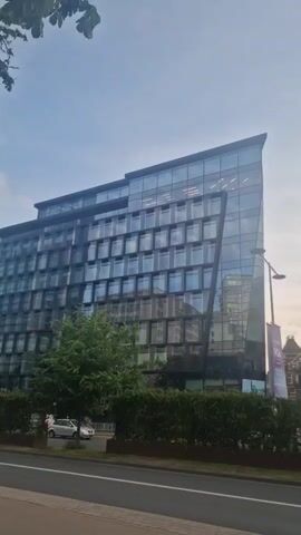The video captures an exterior view of a modern office building with a glass facade. A tree is visible in front of the building. A bus is driving past on the road in front of the building. The sky is cloudy. The camera is positioned at a street level looking up at the building.