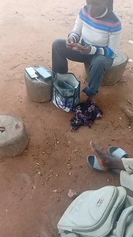 A person with dark skin and braided hair, wearing a striped long-sleeved shirt and dark pants, sits on a wooden stump outdoors. They hold an object in their hands and appear to be speaking. To their left, on another wooden stump, are two smartphones and a bag. A dark floral patterned fabric lies on the ground near the bag. In the foreground, another person's legs and feet are visible, wearing sandals. A light-colored backpack is also present in the foreground. The ground is dirt and appears to be an outdoor setting. The video shifts to show a different person, a man wearing a cap, a light blue denim jacket over a green shirt, and light-colored pants, sitting on what appears to be a large rock. They are also holding something in their hands and seem to be speaking. A light-colored backpack is near them on the ground. The environment is consistent with the previous shot, an outdoor, dirt-covered area.