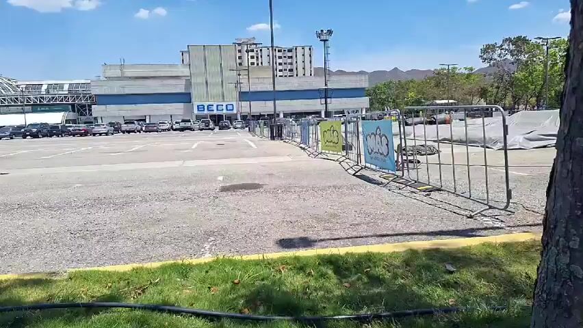 The video shows a parking lot on a sunny day. The parking lot is mostly empty.  Buildings can be seen in the background.  There are a few cars parked.  A patch of grass is in the foreground with a black hose. Some metal barriers with signs on them are visible.