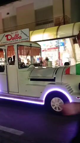 A white vehicle with purple underlighting is parked on a street at night. The vehicle appears to be an ice cream cart or similar food vendor. A sign on the vehicle reads "Dalba" and includes a website address and phone number. People are walking past the vehicle, some appearing to be customers. The street is paved and has yellow lines indicating traffic lanes. Buildings with lights are visible in the background.
