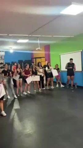 A man stands in front of a line of women, applauding, in what appears to be a gym or fitness studio. The women are holding up certificates. The gym has walls painted in bright colors - green, blue, pink, and yellow. There is a projector screen to the right of the man. The floor is black. The man walks toward the right, still clapping. As he moves, gym equipment comes into view – a barbell and weights. He gives a woman a high-five. A neon sign reading "INTERVAL" is partially visible in the background.
