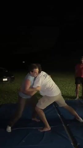 The video shows two young men wrestling on a blue mat outdoors at night. The setting appears to be a grassy field, with a dark sky overhead indicating it is nighttime. A car is visible in the background to the left, and another person, a young man in a red t-shirt and black shorts, stands watching to the right. The wrestling appears to be playful, with both participants smiling and engaged. The mat is segmented, suggesting it might be a portable wrestling or gymnastics mat. The ground is illuminated by artificial light sources not visible in the frame, casting shadows. One of the wrestlers wears a blue t-shirt and brown pants, while the other wears a white t-shirt and khaki shorts. The overall atmosphere seems to be one of friendly competition and recreation.