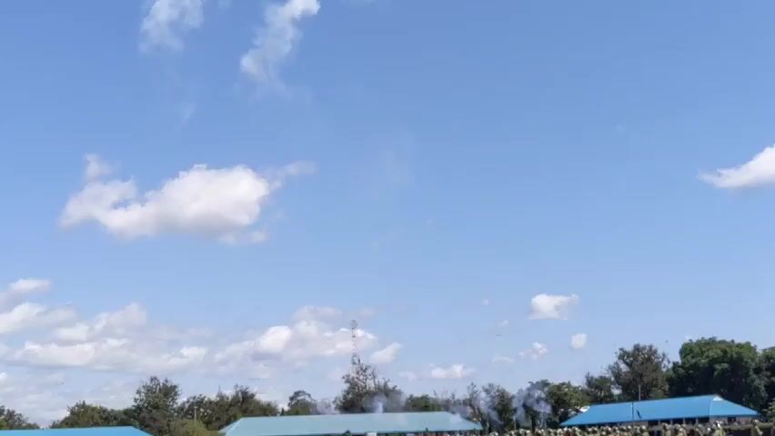 Here's a forensic description of the video:

The video appears to show an outdoor scene, possibly a ceremony or celebration. The sky is bright blue with scattered white clouds. In the foreground, there are some trees and buildings with blue roofs. 

The most notable event is the firing of some type of explosive devices into the air. These explosions leave small, white puffs of smoke against the blue sky. The sound of the explosions is captured in the audio. It looks like multiple explosions occur in quick succession. The purpose of the explosions is unclear, but given the context of a possible ceremony or celebration, they may be intended as a festive display. A group of people in the background seem to be watching the events in the sky.