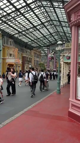 The video captures a busy indoor street scene. Numerous people are walking along a wide pavement with a red border. The street is covered by a large, transparent, grid-like roof supported by a metal structure. Buildings with colorful facades line both sides of the street, resembling a theme park or a shopping mall. The architecture features multiple stories with windows and decorative elements. Several individuals are wearing school uniforms, consisting of white shirts and dark pants or skirts, and many carry backpacks. One person is pushing a wheelchair. The lighting appears to be natural light filtering through the roof, supplemented by artificial street lamps with hanging flower baskets. The overall atmosphere suggests a public area, possibly a promenade or an entertainment district.