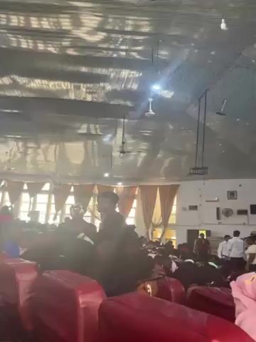 The video shows the interior of a large room with many people sitting in rows of red seats. The room has a high ceiling with a patterned covering and several ceiling fans. There are also pendant lights hanging from the ceiling. Natural light streams in through the windows, which are covered with orange and white curtains. At the front of the room, a stage area is visible with people standing and moving around. Some individuals are wearing white shirts. Some people in the seats are looking around and interacting with each other. The overall atmosphere appears to be busy and populated, possibly an event or gathering.
