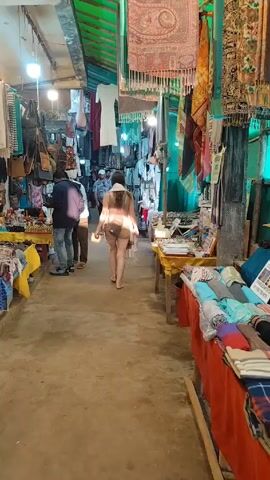 The video shows a narrow market aisle with numerous stalls on both sides displaying items such as clothing, bags, jewelry, and souvenirs. People are seen walking through the aisle, some stopping to browse the merchandise. One woman, wearing a hat and short shorts, is walking away from the camera, carrying a plastic bag. The market area is covered by a green roof and decorated with hanging textiles and lights. The floor is a worn concrete surface.