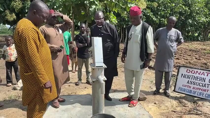 A group of men and a few boys are gathered around a water well in a rural, outdoor setting. The well has a hand pump mechanism. A sign in the background reads "DONATED BY NORTHERN CHRISTIAN ASSOCIATION INC. WITH SUPPORT FROM FOUNTAIN OF LIFE CHURCH LAGOS." The people are dressed in various traditional African attire and modern clothing. One man in a yellow patterned garment on the far left is pointing at the well. Another man in a white vest and cream pants is operating the pump. The ground appears to be dirt and there are trees and vegetation in the background, suggesting a dry, agricultural area. The lighting indicates it is daytime.