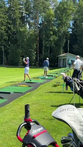 The video shows a golf driving range on a sunny day. Several people are practicing their golf swings on individual mats. In the foreground, a red golf push cart is visible. In the middle ground, two golfers are actively taking swings, with a line of golf balls behind each. Further back, other golfers are lined up, and golf bags are placed by the mats. A small building with a white exterior and a green roof is situated at the far end of the driving range. The area is surrounded by a dense forest of tall green trees.