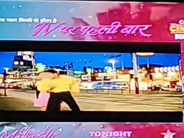 This video shows a scene of a couple dancing on a street at night. The man is wearing a yellow shirt and black pants, and the woman is wearing a pink skirt and a black top. The street is lit up with colorful lights, and there are buildings in the background. The couple appears to be enjoying themselves, and they are dancing in a playful manner. The video also includes some text at the top, which appears to be in Hindi, and translates to "Now love happens for the first time on TV."
