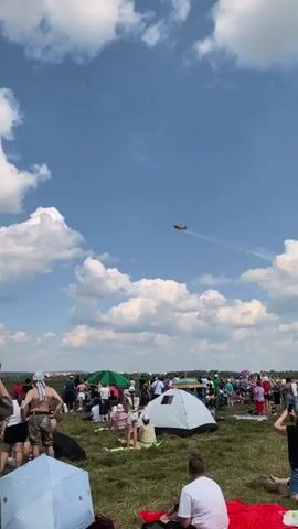 This is a vertical video taken at an outdoor event, likely an airshow, on a sunny day with scattered clouds. A crowd of people, dressed in casual summer clothing, are gathered on a grassy field, many sitting on blankets or chairs. Some individuals are holding up phones, presumably to record or photograph the events. Several tents and umbrellas are visible among the crowd. In the sky, an airplane is performing aerial maneuvers, leaving a smoke trail behind it. The plane appears to be a small, single-engine aircraft. Another, smaller plane is visible in the distance. The overall atmosphere seems festive and engaged, with spectators focused on the aerial display.