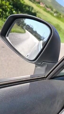 The video shows a view from inside a car driving on a paved road. The perspective is from the passenger side, looking out the window and into the side mirror. The side mirror shows a reflection of the road ahead, which appears to be uneven and cracked. The passenger window frame is visible in the foreground. Outside the car, there are some green bushes and trees, as well as a few houses in the distance. The sky is blue with a few scattered white clouds. The sun is shining, casting shadows. The houses appear to be residential, with red-tiled roofs. One house has a balcony. There are also utility poles and wires visible. The overall impression is of a rural or suburban setting on a clear, sunny day.