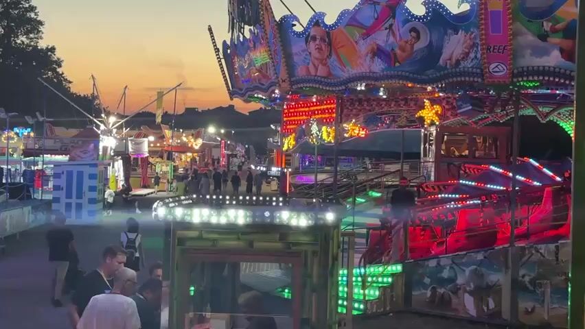 This media file captures a vibrant amusement park scene at dusk in Riccione, Italy. The main subject is a large, brightly lit carnival ride adorned with colorful graphics depicting surfing and ocean themes, including waves, dolphins, and people on surfboards. The ride itself features multiple levels and is illuminated with a dazzling array of lights, including green, pink, purple, and red LEDs, creating a festive and energetic atmosphere.

In the foreground, several people are visible, some appearing to be park-goers and others possibly staff. A few individuals are gathered near the entrance of a structure that seems to be part of the ride or an adjacent attraction, with one person in a white shirt. Further back, more people are walking through the park, suggesting a lively evening crowd.

The setting is an amusement park with other rides and attractions visible in the background. The sky is a soft gradient of orange and pink, indicating sunset. The overall mood is one of excitement and enjoyment, characteristic of a summer evening at a fairground. The lighting, especially the flashing and colorful LEDs, is a prominent detail, highlighting the dynamic nature of the amusement park. There are no discernible weather conditions other than the twilight sky. No specific text is clearly readable on the ride or in the environment, though there is a sign on the ride that seems to say "REEF".