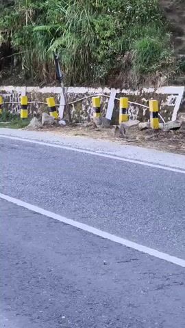 The video shows a road curving to the left. The road has a white line down the middle and a rough, cracked surface. On the right side of the road, there is a steep embankment covered in trees and vegetation. Yellow and black striped bollards are spaced along the top of a low stone wall that runs parallel to the road. A tall pole with red and white bands stands near the side of the road. The scene appears to be in a rural or mountainous area.