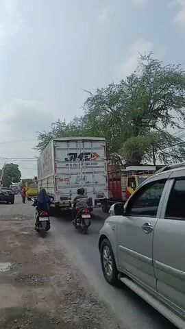 The video shows a traffic jam on a paved road under a cloudy sky. Various vehicles, including cars, motorcycles, and trucks, are stopped in traffic. A large white truck with "JNE" branding is visible, along with a silver SUV and several motorcycles. People are riding the motorcycles, some wearing helmets. A large tree with green foliage is on the right side of the road, and there are buildings and more trees in the background. The road surface appears rough in places.