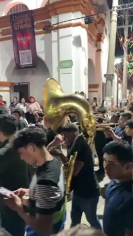 The video shows a nighttime street scene with a brass band playing amidst a crowd of people. The camera is positioned within the crowd, looking slightly upwards.

The band is composed of several musicians playing brass instruments, including a tuba. They are surrounded by people who appear to be enjoying the music, many of whom are holding drinks. The crowd is diverse in age and attire. The street is lined with buildings, and colorful lights are strung overhead, adding to the festive atmosphere. A banner hangs on a building on the left side of the frame. The overall impression is one of a vibrant street party.