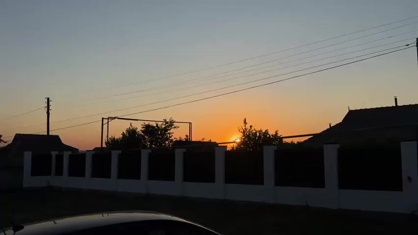 The video shows a sunset over a rural or suburban area. A dark fence with white posts runs horizontally across the lower portion of the frame. Behind the fence, silhouetted trees and the tops of houses are visible against the orange and blue sky. The sun is low on the horizon, partially obscured by trees, casting a warm glow. Power lines are strung across the sky. In the foreground, the silhouette of a car is visible. The scene appears to be still, with no people or discernible activity.