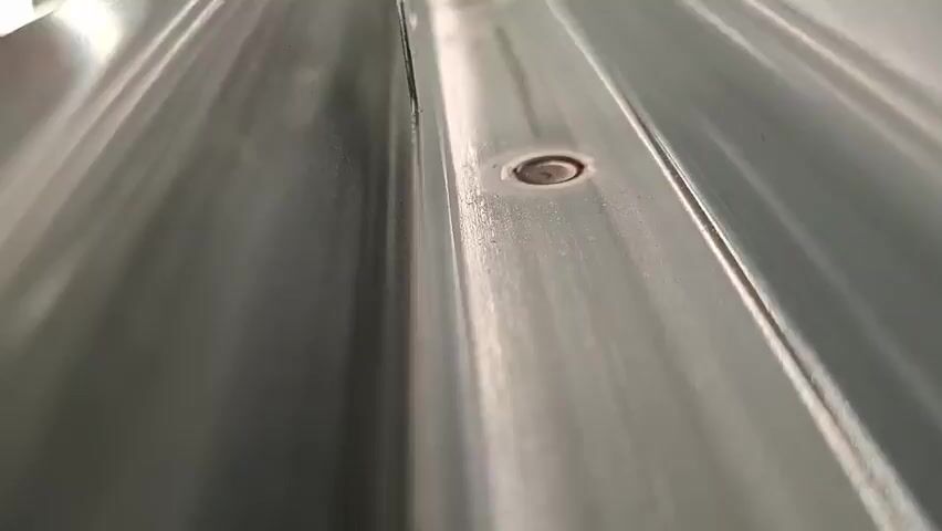 The video shows a close-up, low-angle, forward-moving shot of a corrugated metal surface. The surface is light gray and appears to be made of aluminum or galvanized steel, with the characteristic raised ridges and channels. Several circular fasteners, likely rivets or screws, are visible along the ridges. The lighting creates reflections and highlights on the metallic surface, emphasizing its texture and shape. The camera movement is smooth and steady, suggesting it is mounted on a stable platform or a drone. The focus is sharp on the metal surface, and the background is blurred, further isolating the texture of the material. There are no people or other objects visible in the frame besides the metal surface and its fasteners. The environment appears to be outdoors, possibly a roof or a wall, given the nature of the material. The time code indicates the duration of the clip.