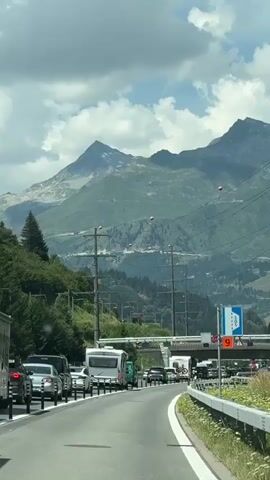 A vertical, first-person perspective shot shows a traffic jam on a highway with a mountain range in the background. The road curves to the right, with several lanes of vehicles including cars, recreational vehicles, and trucks. There are power lines and poles running overhead. The sky is partly cloudy. The mountains are covered in trees and have rocky peaks. Orange markers are visible on the power lines. A highway sign indicating a speed limit of 60 is visible.