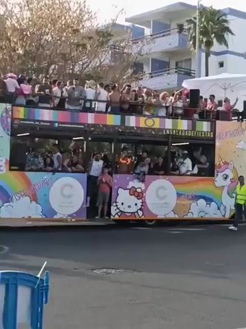 The video captures a vibrant LGBTQ+ pride parade. A double-decker bus decorated with "ENSALADADEFIESTAS" and images of Hello Kitty and a unicorn moves along a street crowded with participants. People on the bus, mostly men, wave and cheer. On the ground, a diverse group of individuals walks alongside the bus, some dancing. A rainbow flag is prominently displayed near a supermarket. The setting appears to be a sunny, outdoor location with buildings and palm trees in the background.