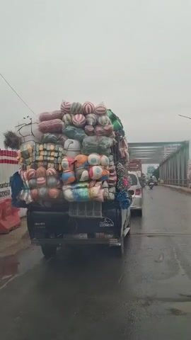 A black pickup truck is overloaded with goods. The cargo is piled high above the truck's cab and consists of many round, colorful items, possibly stuffed animals or balls, along with other bagged and wrapped items. The truck is on a wet road, suggesting it may have rained recently. In the background, a metal bridge spans across a body of water. Other vehicles, including a white car and motorcycles, are also on the road. One person on a red scooter is visible on the left side of the frame, and a person on a motorcycle is seen further down the road. The sky is overcast and the overall atmosphere appears to be one of everyday travel.