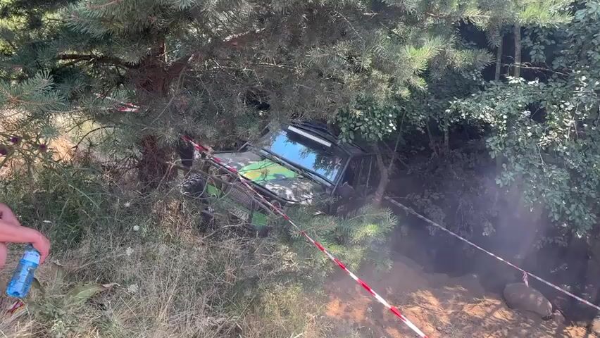 A black off-road vehicle with green accents is stuck on its side, entangled in tree branches. Red and white caution tape is visible, strung between trees and across the muddy ground. A person's arm, holding a blue water bottle, is visible in the lower left corner of the frame. The vehicle appears to be in a wooded, outdoor area with dense foliage. The overall scene suggests a recovery or rescue operation in progress after the vehicle became stuck.
