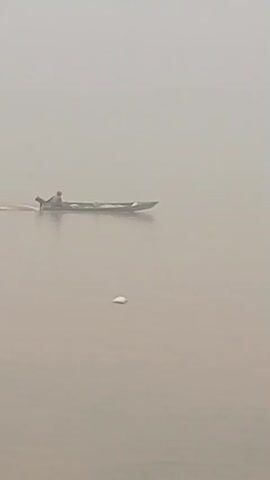 A person is in a long boat, which is moving through murky water. The boat has a motor attached to the back. There are two small white objects in the water, one closer to the boat and one further away. The water is calm, and there is a hazy, light-colored sky or fog in the background.