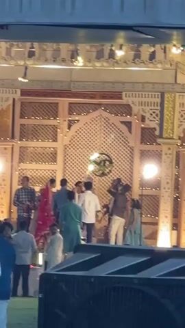 The video shows an outdoor wedding celebration. A bride in a red outfit and a groom in white attire stand on a stage decorated with intricately designed wooden panels and illuminated by several stage lights. A group of people, some dressed in formal attire, surround the couple, seemingly congratulating them and capturing the moment with cameras. The stage is set against a backdrop that appears to be a structure covered with a canopy or awning. In the foreground, parts of sound equipment are visible, and other guests are seen milling around, adding to the celebratory atmosphere.