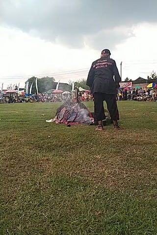 Here's a concise description of the video:

The video shows a man in a black jacket and pants kneeling on a grassy field near what appears to be a burning or smoking object. The man seems to be interacting with the object, possibly adjusting it or watching it.  A crowd of people and some structures are visible in the background, suggesting an event or gathering is taking place. The sky is overcast. The video is vertically oriented.