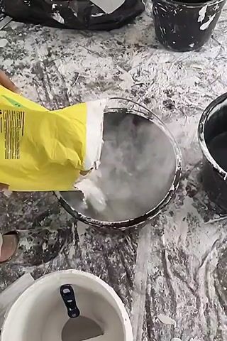 A close-up shot shows a yellow bag being tipped over a clear bowl. White powder pours from the bag into the bowl, which already contains some liquid and powder. The bowl is on a surface covered in a white tarp, and there are other buckets and tools visible in the background, also covered in white powder. The scene appears to be a workspace where some kind of material is being mixed.
