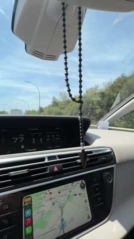 The video shows the interior of a car, likely during daytime, as seen from the passenger's perspective. A set of black prayer beads hangs from the rearview mirror, swaying slightly. The dashboard displays a digital speedometer showing 77 km/h, and the navigation system is active on the central screen, displaying a map. Outside the car, trees and a bright sky are visible, suggesting the vehicle is on a road, possibly a highway given the signage in the distance. The car appears to be in motion. There are no people clearly visible in the frame.