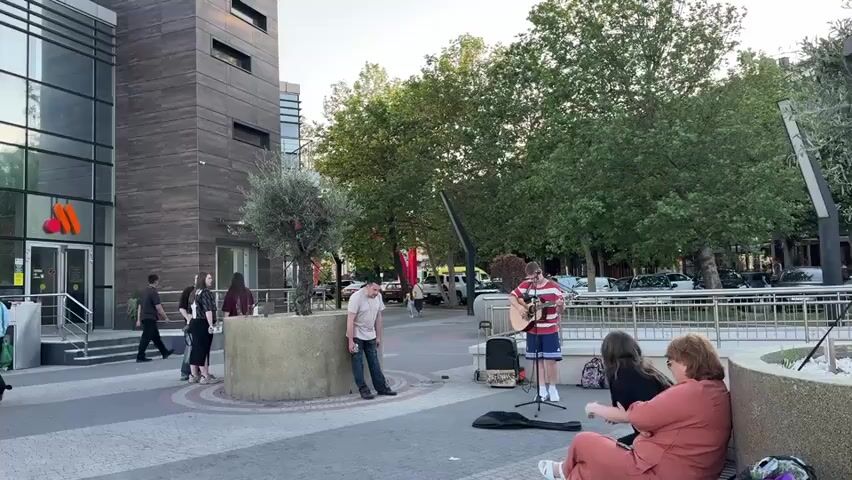 The video shows a street scene with several people. In the foreground, a man is playing a guitar and singing. Two women are sitting on a bench nearby, listening to him. In the background, there is a modern building with large glass windows. Other people are walking around on the sidewalk. The scene appears to be a public space, possibly a plaza or park, with trees and benches. The lighting suggests it is daytime, with a clear sky.