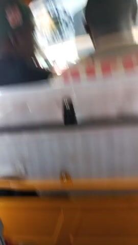 The video shows the inside of a moving auto rickshaw, also known as a "tuk-tuk."  Two passengers are visible, one wearing a green baseball cap and a black shirt, and the other with a white shirt with "DIESEL" written on it. The interior of the tuk-tuk is simple, with a seat partially covered in plastic wrap. Outside, the road is visible, with other vehicles and people passing by, and there is a glare from the sun. The view from the tuk-tuk shows the open road ahead, with houses on the side. The perspective is from the back seat of the vehicle.